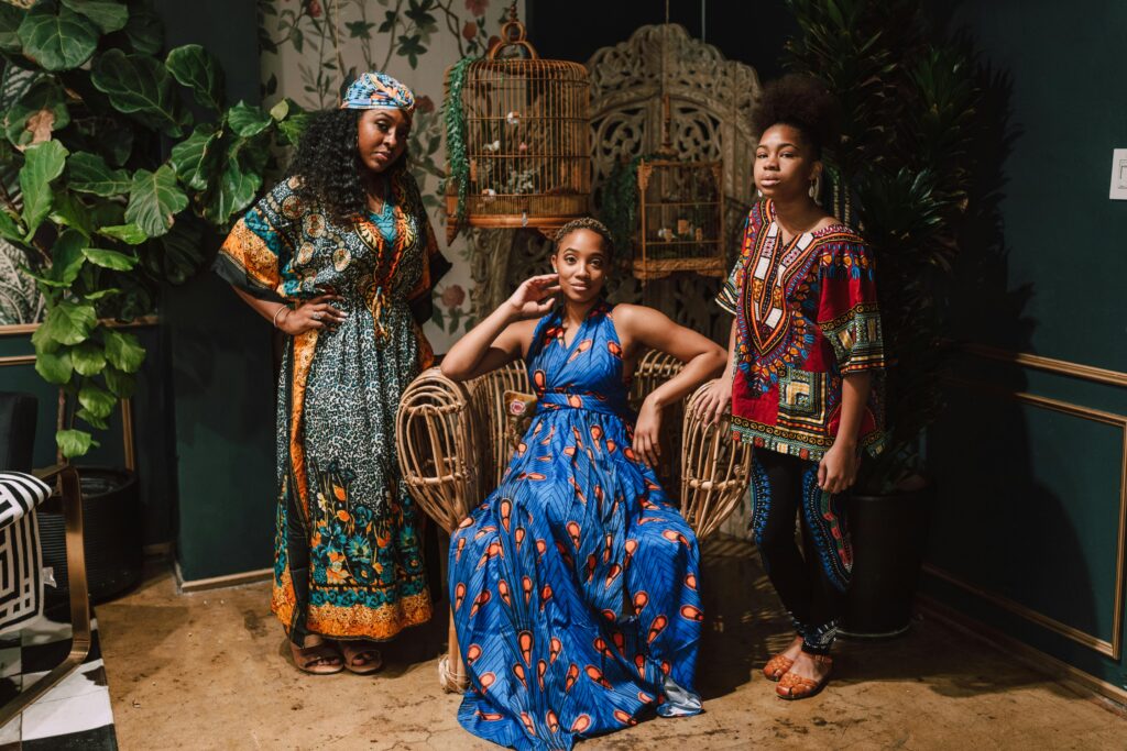 Three models in traditional African attire posing with style indoors surrounded by plants and birdcages.